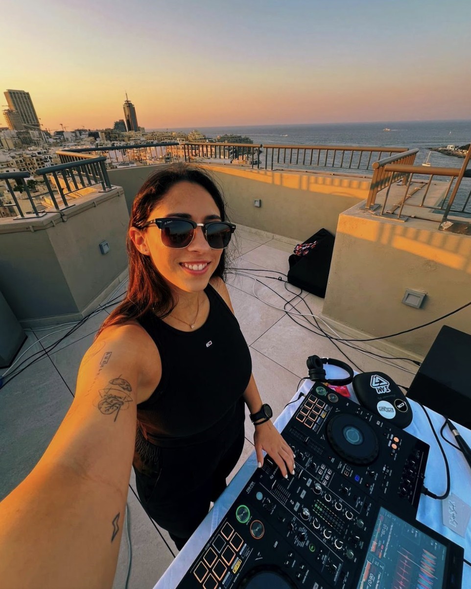 Woman DJ in black tank top smiling on a rooftop at sunset with city and ocean views.