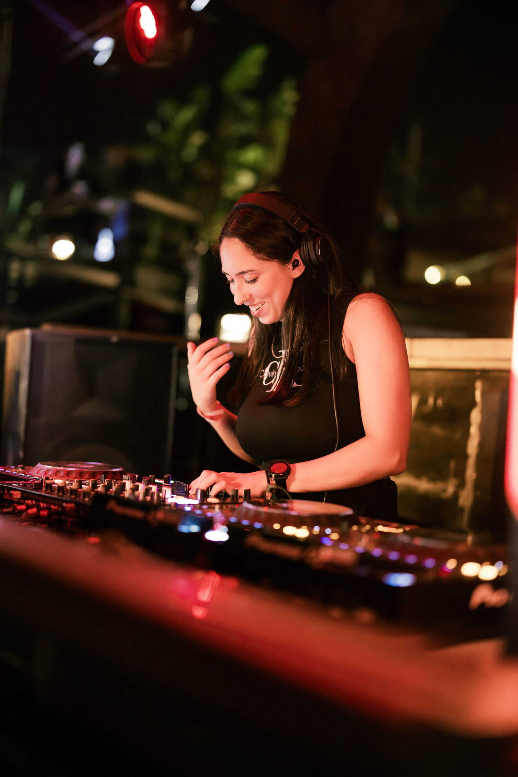 DJ in black top smiling behind a mixing console under vibrant club lights.