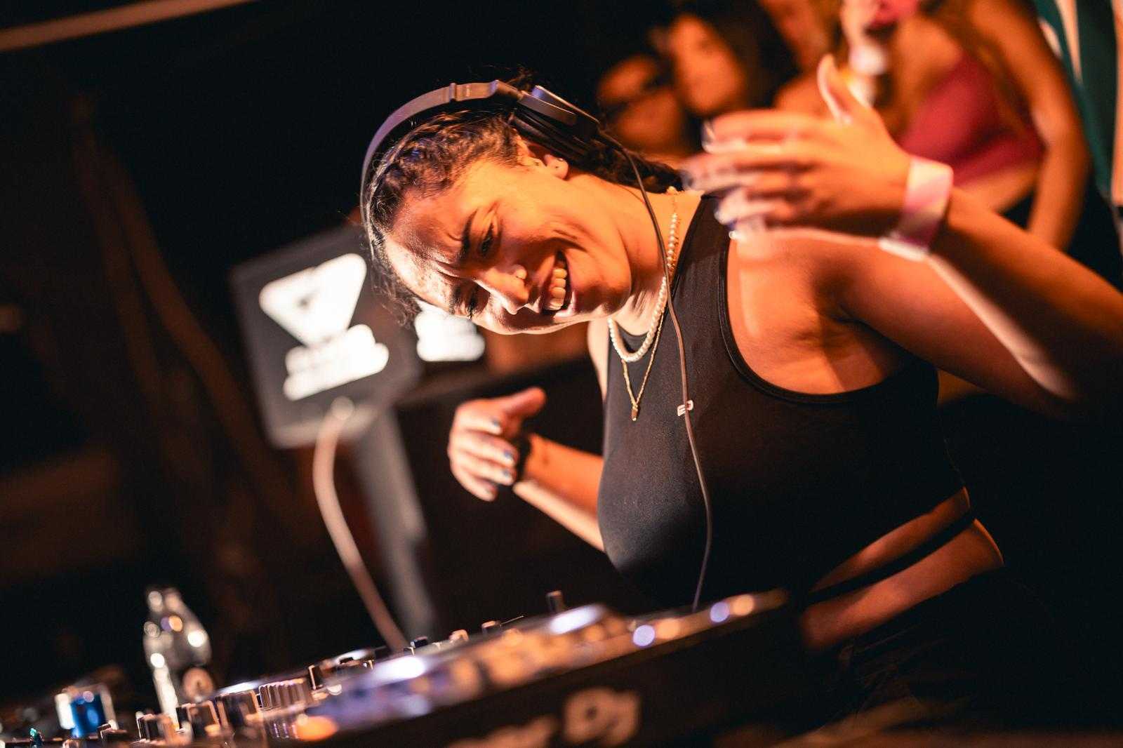 Club DJ with headphones, smiling and gesturing, lively crowd surrounding.