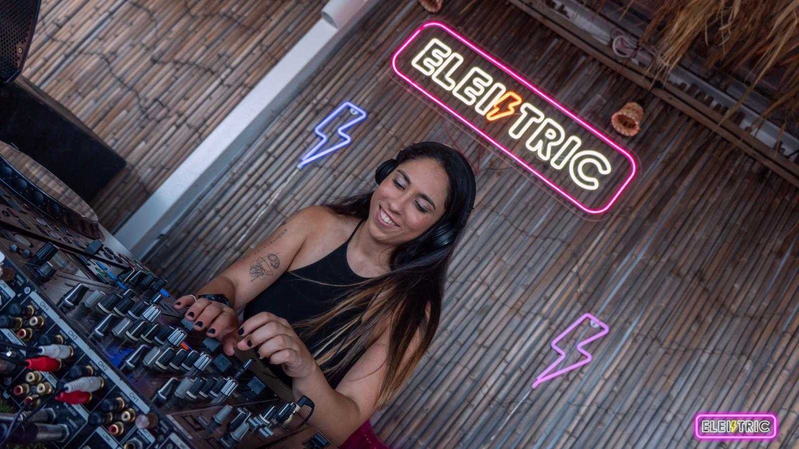 DJ smiling while mixing music, wooden wall with neon "Electric" sign behind.