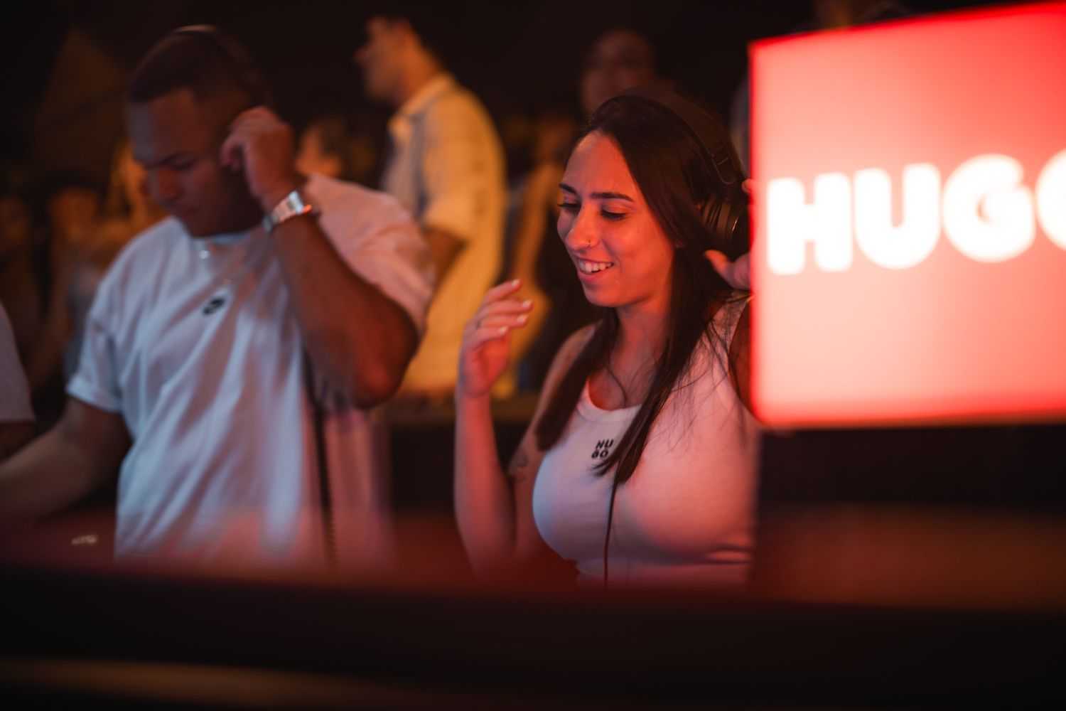 Two DJs at a booth, man focused, woman smiling, "HUGO" sign visible.