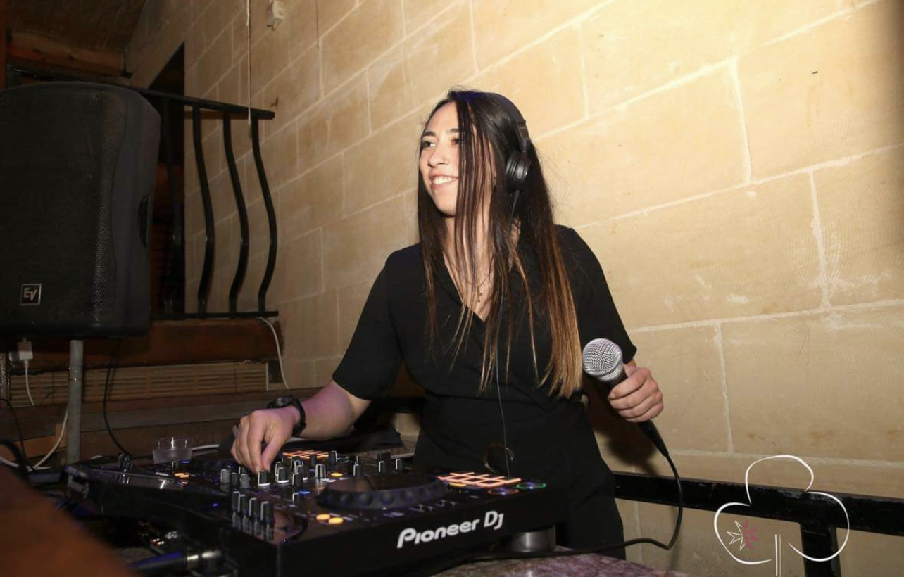 Female DJ in black dress holding a mic and mixing music on stage.