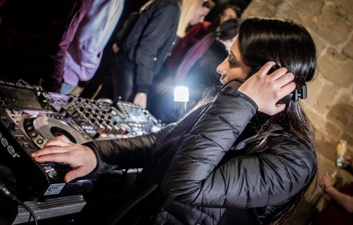 DJ in a black jacket mixing music, focused on equipment, lively crowd in the background.