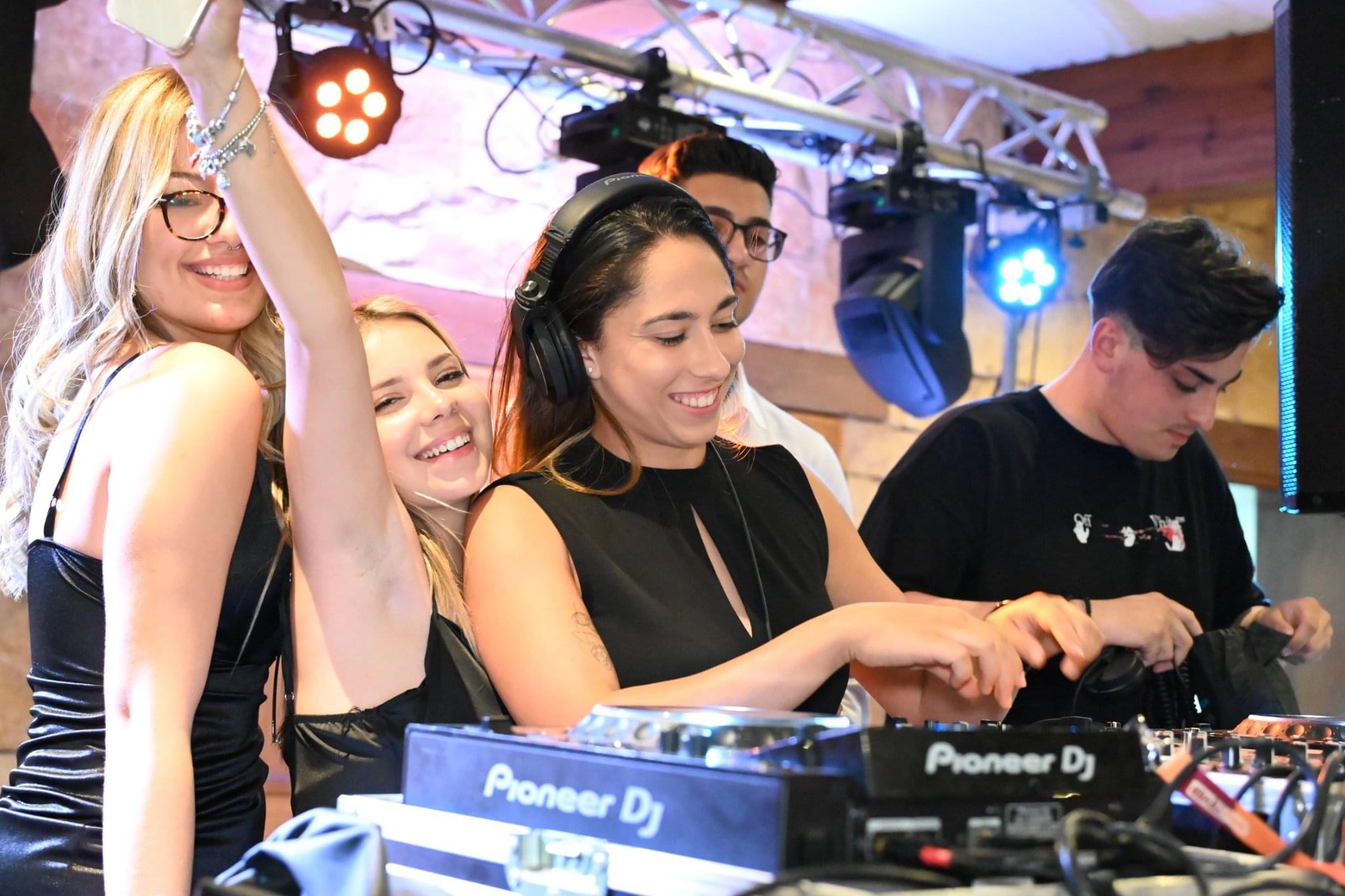 Party scene with a female DJ in the center mixing tracks, two women dancing beside her.