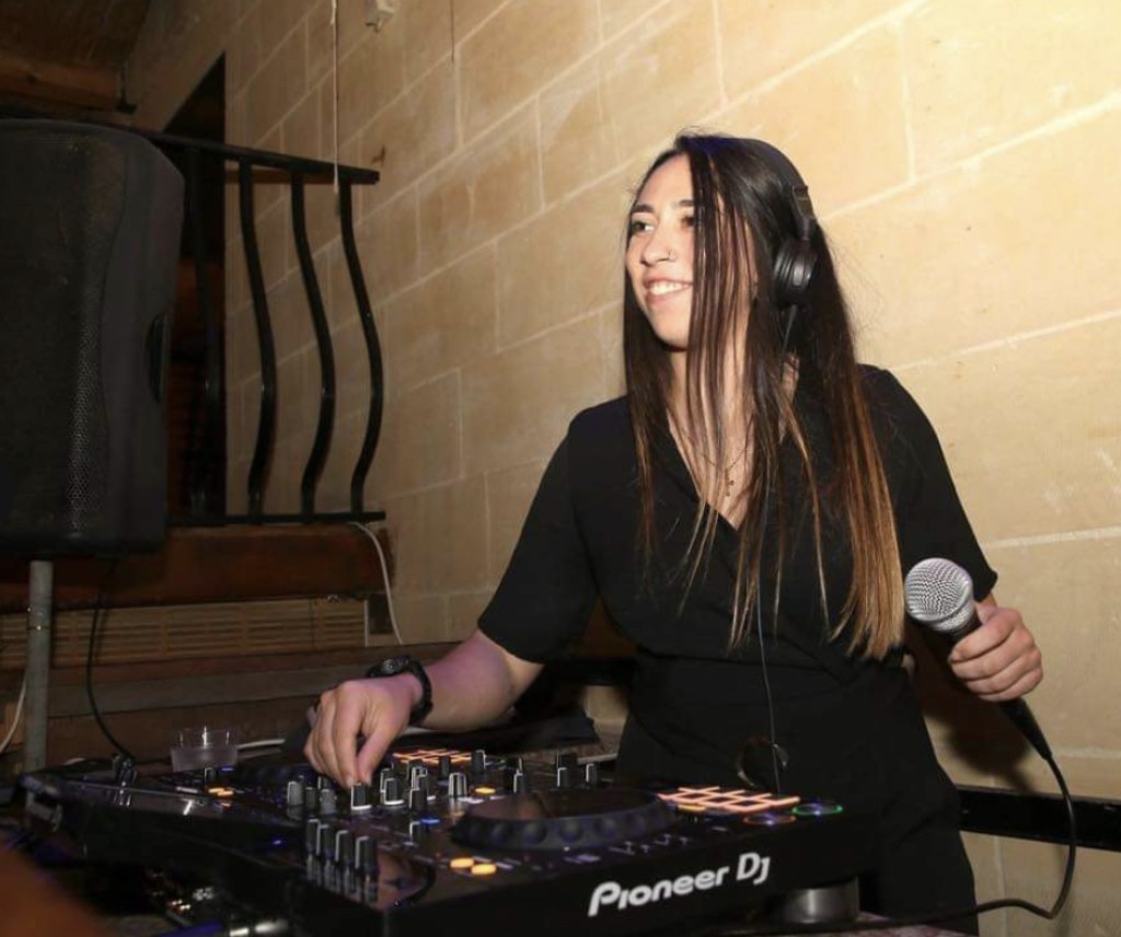 DJ smiling with mic, adjusting music controls in a dimly lit room.