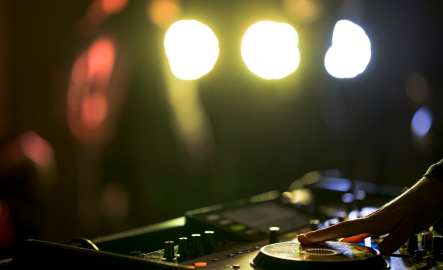 Hand on DJ mixer in dim lighting, bright stage lights in the background.