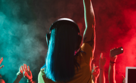 DJ with blue hair and headphones raising a hand, vibrant lighting and lively crowd.