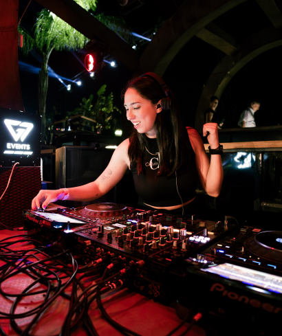 DJ with long hair smiling while mixing music under an arched structure.