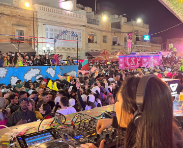 Outdoor DJ at a festive event, colorful floats and decorations, lively crowd dancing.