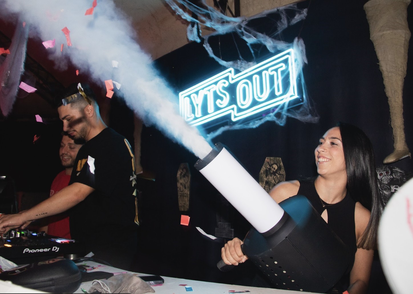 Woman holding a fog machine at a party, DJ and neon "LYTS OUT" sign behind.