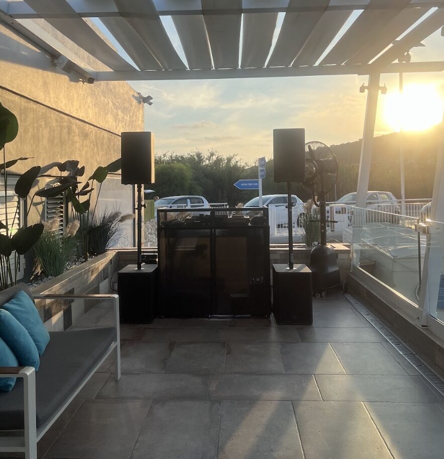 Outdoor patio DJ setup under a pergola at sunset, with hills and parked cars visible.